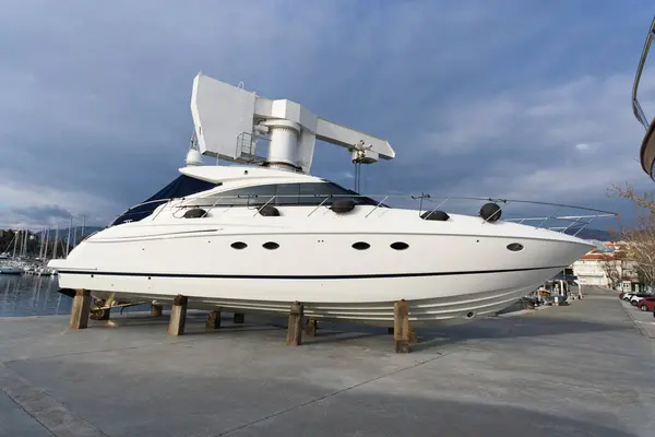 Parlak beyaz bir yat, ahşap desteklerle yükseltilmiş, çarpıcı bir gökyüzünün altında beton bir rıhtımda duruyor, modern tasarım ve radar ekipmanlarını sergiliyor..