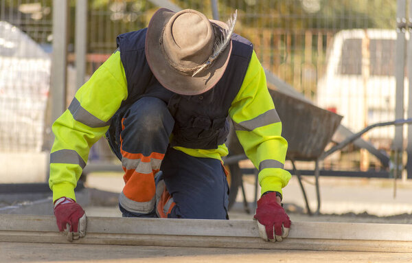 A paver (specialist - foreman) with a leather hat (with a feather) on his head and wearing reflective protective work clothes, uses an aluminum "board" to level ("pull down") the smallest fraction of the subgrade for paving. 