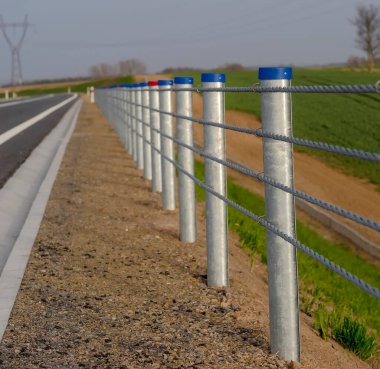 Sıralarda bariyerler olan bir yol. Baharda yolun kenarındaki çeliklerden ve direklerden yapılmış bir korkuluk. Bahar öğleden sonra. Resimli ve kullanışlı, çelik halatlardan yapılmış, tarlalardan gelen hayvanlara karşı yolu koruyor.. 