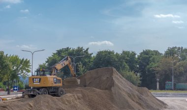 Polonya Ostrowiec Swietokrzyski 22 Eylül 2023, saat 11: 51. Bir CAT tekerlekli kazıcı bir çakıl yığınının içinde hareket ediyor. Şehirdeki ağaçların arasında mavi gökyüzünün altındaki bir yol inşaatında büyük tekerlekli bir kazıcı..  