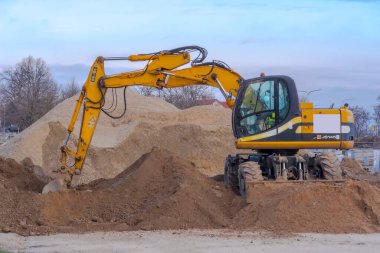 Polonya Ostrowiec Swietokrzyski 19 Mart 2024 saat 17: 07 'de. JCB kazıcısı bir çakıl yığınının üzerindeki inşaat alanında. Yol inşaatı - büyük çakıl yığını ve kazıcı bir kamyonun yüklenmesini bekliyor, bir bahar akşamı mavi gökyüzünün altında..  