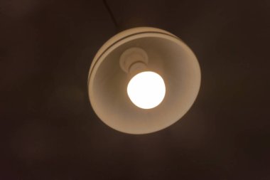 Beyaz arka planı olan bir ampul. Karanlık bir odada parlayan bir lamba. Bir abajurlu tavan lambası ve parlayan bir LED lambası..  