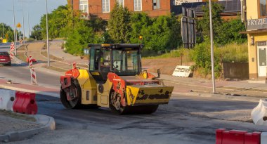  Poland Ostrowiec Swietokrzyski June 26, 2024 at 18:43. The large Dynapac road roller compacts the asphalt layer. A road construction site illuminated by the setting sun. A large road machine in action, an old historic brick house in the background . clipart