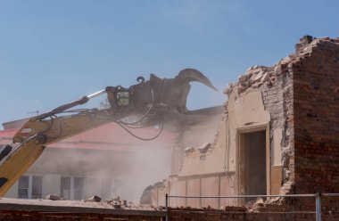  Kırmızı tuğlalı bir binanın son derece tozlu yıkım alanı. Eski binayı dekonstrüksiyonlu bir kazıcı. Şehirdeki bir binanın yıkım alanı - bir kazıcı çelik çeneli tuğla bir duvarı 