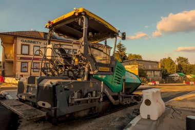 Poland Ostrowiec Swietokrzyski May 28, 2024 16:51. Asphalt paving of the renovated road   VGELE asphalt paving machine on a road under renovation .The machine features a prominent canopy and is positioned on freshly laid asphalt.Two-story building . clipart