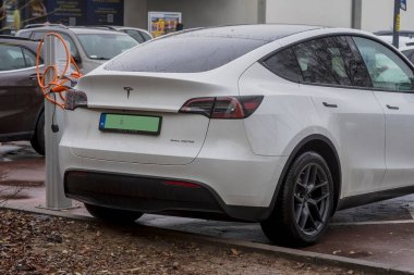 Polonya Ostrowiec Swietokrzyski 28 Kasım 2024: 36. Yağmurlu bir günde şarj istasyonuna park etmiş beyaz Tesla elektrikli arabası pillerini şarj ediyor. Araç turuncu şarj kablosu ile bağlanmış. Arka planda bir işaret görünüyor.