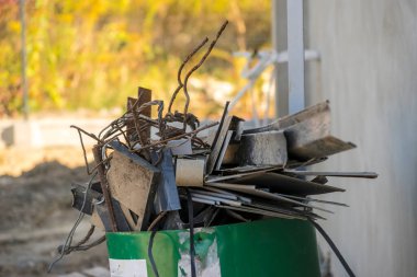 Yeşil bir varil, inşaat alanında çelik çubuklar, levhalar ve plakalar da dahil olmak üzere çeşitli metal kırıntılarla doludur. Metal parçalar paslanmış ve düzensiz bir şekilde namlunun kenarından çıkmış. Arkaplan bulanık yaprakları gösterir.