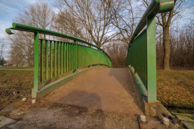 Dikey korkulukları olan küçük yeşil çelik bir köprü dar bir kanal boyunca park ortamında uzanıyor. Köprü döşemesi desenli metal gibi görünüyor, hafif kavisli. Etrafında ağaçlar ve otlar vardır..  