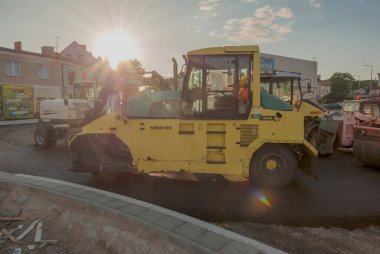 Polonya Ostrowiec Swietokrzyski 5 Haziran 2024: 46. Batan güneşin altında bir yol inşaatında inşaat makineleri. Güneş, etrafa dağılmış bulutlarla bölgeyi aydınlatan sıcak bir parıltı saçar. İşçiler makinelerin içinde görülebilir..