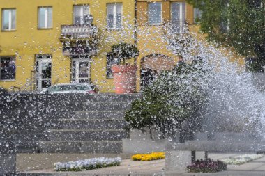 Bir bahar günü, tarihi pazar meydanındaki şehir çeşmesinden su püskürtüleri. Çeşmeden çağlayan su püskürtüleri, tarihi bir pazar meydanının zeminine dinamik bir sprey püskürtüyor. Arka planda sarı bir bina var. . 