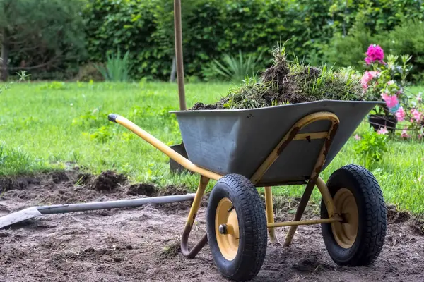 Bahçedeki toprak işleri (el arabası, kürek, vs.) Çiçek tarhını düzenlerken. Toprak ve çimen yığınlarıyla dolu gri metal bir el arabasıyla bir bahçe sahnesi. El arabasının sarı tekerlekleri var ve çıplak bir toprak parçasına yerleştirilmiş.. 