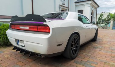 Polonya Ostrowiec Swietokrzyski 11 Temmuz, 2025 'te Beyaz DODGE CHALLENGER' in yanında tarihi bir kilisenin yanında tuğla bir yola park edilmiştir. Arabanın arka tarafında açık renkli duvarları olan tarihi bir kilisenin yanındaki renkli camlar var. .