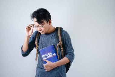 Asyalı genç bir erkek öğrenci çanta takıyor, kitap okuyor, gülümsüyor ve gözlük takıyor. İzole edilmiş beyaz arkaplan