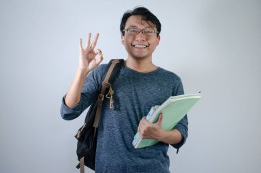 Asyalı genç erkek öğrenci çanta takıyor, elinde kitap tutuyor gülümsüyor ve mutlu bir ifade takınıyor. İzole edilmiş beyaz arkaplanı olan el işareti hareketi