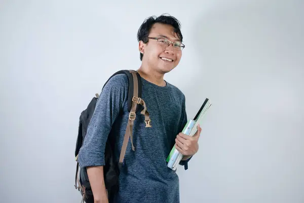 Asyalı genç erkek öğrenci çanta giyiyor ve kitap taşıyor. Gülümsüyor ve mutlu bir ifadeyle izole edilmiş beyaz bir altyapısı var.