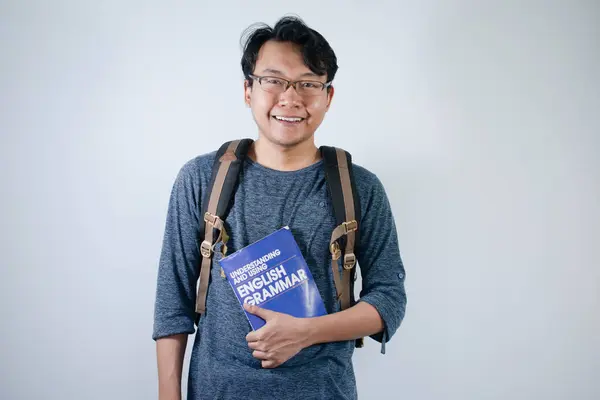 Asyalı genç erkek öğrenci çanta giyiyor ve kitap taşıyor. Gülümsüyor ve mutlu bir ifadeyle izole edilmiş beyaz bir altyapısı var.