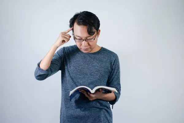 Asyalı genç erkek kitap okuyor ve jest yapıyor. Gözlüklü Asyalı erkek. Gri tişörtlü, izole beyaz arka planı var.