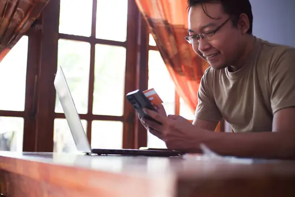 Gömlekli Asyalı bir erkek oturma odasında kredi kartı akıllı telefonu ve dizüstü bilgisayarıyla online işlem yapıyor. Evde çalışan serbest çalışan bir işçi.