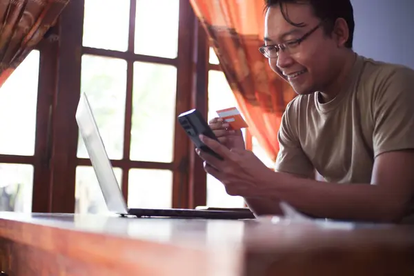 Gömlekli Asyalı bir erkek oturma odasında kredi kartı akıllı telefonu ve dizüstü bilgisayarıyla online işlem yapıyor. Evde çalışan serbest çalışan bir işçi.