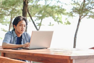 Ciddi Asyalı genç iş adamı dışarıda dizüstü bilgisayarla çalışıyor. Uzaktan kumanda ve serbest çalışma kavramı. Sıradan mavi elbiseler giyen başarılı genç adam.