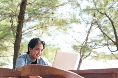 Mutlu Asyalı genç iş adamı dışarıda dizüstü bilgisayarla çalışıyor. Uzaktan kumanda ve serbest çalışma kavramı. Sıradan mavi elbiseler giyen başarılı genç adam.