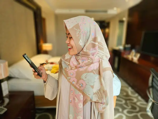 Mutlu Asyalı Müslüman kadın tesettürlü, mesaj okurken gülümseyen ya da arka planda telefonla sohbet eden, telefonla iletişim kuran, modern küresel internet sosyal medya kavramı