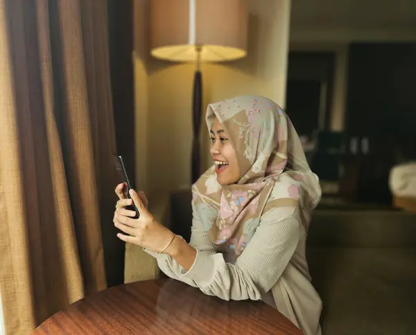 Mutlu Asyalı Müslüman kadın tesettürlü, mesaj okurken gülümseyen ya da arka planda telefonla sohbet eden, telefonla iletişim kuran, modern küresel internet sosyal medya kavramı