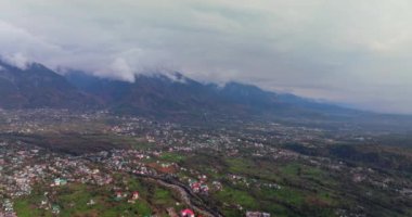 Dharamshala şehrinin hava manzarası, bulutlarla kaplı Dhauladhar dağları, renkli evler ve çerçevede görünen yeşil çiftlikler, 4k video