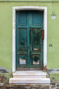 Bir çift eski ve aşınmış çift ahşap kapılı altı işlemeli dikdörtgen. Boya soyuluyor ve çatlıyor. Sağ tarafında paslı bir dahili telefon kutusu var. Her orta panelde iki metal kapı kolu var..