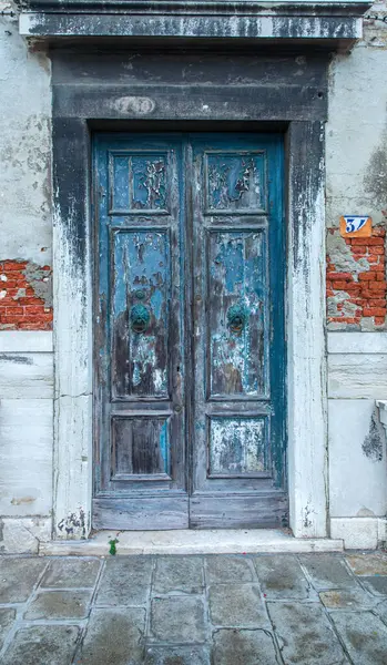 This is an image of a pair of faded green double doors. The door has six panels with two metal doorknobs. The paint is peeling and cracked. The door handles are metal rings attached to a lions head positioned in the middle of the center panel.
