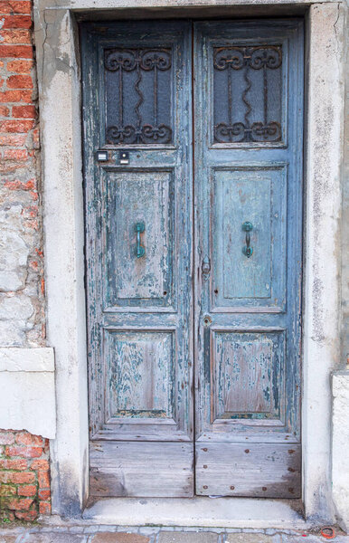 This is an image of a pair of faded green double entrance doors. The door features two metal handles, a keyhole and a doorbell. Each door has three square recessed panels, the top panels are glass and are protected by decorative metalwork.