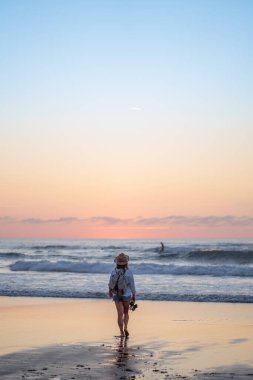 Beyaz gömlekli ve şapkalı bir adam gün batımında sahilde yürüyor..