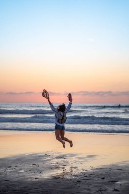 Gün batımında sahilde atlayan sörf tahtası olan genç bir kadın. Güzel gün batımı.