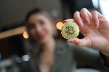 Güzel Asyalı iş kadını, elinde bir eliyle altın bir bitcoin tutarken önünde bir yığın bozuk para, hesap makinesi ve siyah masada bir tablet, altın bitcoin üzerine odaklanıyor. Yüksek