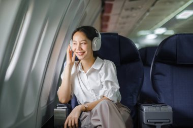 Uçakta oturan ve akıllı telefon kullanan Asyalı kadın uçak koltuğu pencere yolculuğu ve teknoloji konseptinin yanında müzik dinliyor. Yüksek kalite fotoğraf