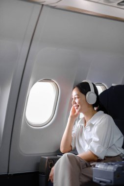 Uçakta oturan ve akıllı telefon kullanan Asyalı kadın uçak koltuğu pencere yolculuğu ve teknoloji konseptinin yanında müzik dinliyor. Yüksek kalite fotoğraf