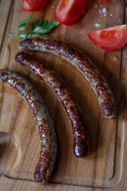 Kızarmış patates dilimleri, taze domates dilimleri ve ahşap bir tahtanın üzerinde bir diş sarımsak ile servis edilen ızgara sosisler..