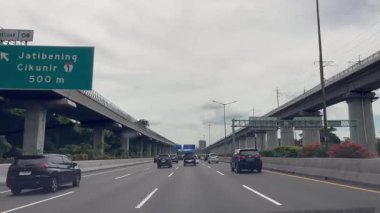 Dashcam View Araba Sürüşlü Şehir Otoyolu. Hareket halindeki araçların, yükseltilmiş tren raylarının ve yakınlardaki yeşilliklerin yer aldığı modern bir şehir otoyolu sahnesi.,