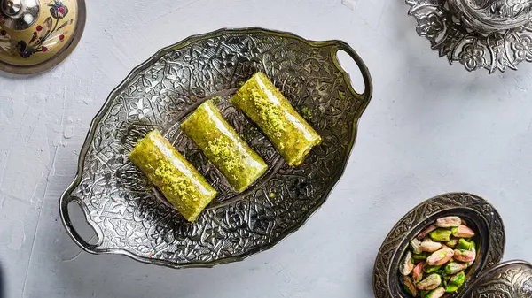 Pieces of oriental sweets with pistachios in a white plate on a white ground surrounded by silver and copper utensils