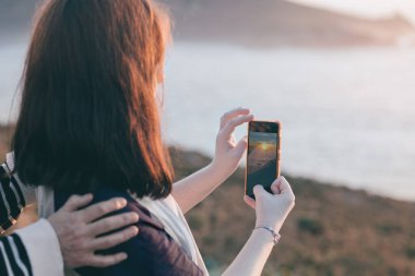 Genç bir kadın gün batımında cep telefonuyla okyanusun resmini çekiyor annesiyle geçirdiği güzel anın tadını çıkarıyor.