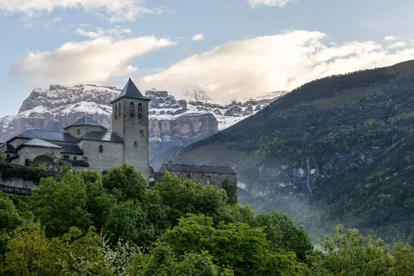 stock image Torla, a charming village nestled in the Aragonese Pyrenees, Spain. The old church surrounded by lush vegetation with the snow-capped mountains in the background and a partly cloudy sky.