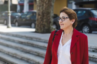 Kırmızı ceketli ve gözlüklü olgun bir kadın şehir meydanında dikilirken hafifçe gülümser ve şehir hayatının samimi bir anını yakalar..