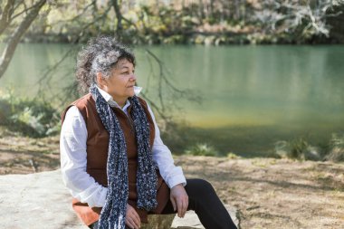 A Cuban woman enjoys a peaceful moment sitting by the tranquil river in Fragas do Eume, Galicia, Spain clipart