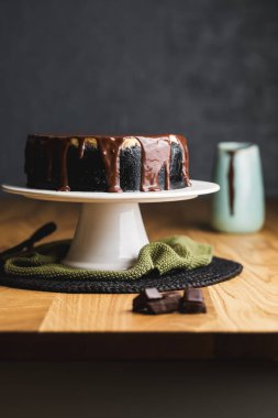 Koyu kurabiye kabuğuyla süslü zarif bir cheesecake, üzerinde zengin çikolata ganache, beyaz pasta standında sade dekorlu..