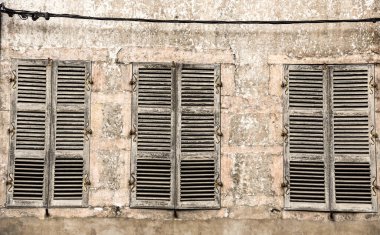 Üç ağır yıpranmış pencere ve kapalı jaluziler eski bir hava duvarının önünde duruyor.