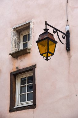 İki küçük pencereli pembe duvar ve sarı camlı klasik bir sokak lambası.