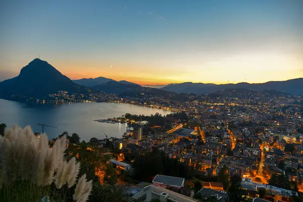 Lugano Gölü 'nün üzerinde gün batımı. Şehrin ışıkları yanıyor.