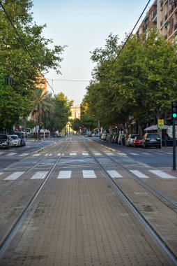Sessiz Valencia caddesi, bir yaz sabahı, tramvay rayları, ağaçlar, park edilmiş arabalar ve yumuşak altın ışıkla