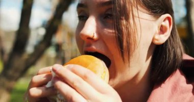 Güzel kız fast food yiyor. Parkta yemek yiyor. Kadınlar gün ışığında sokak yemeklerinden hoşlanır. Yüksek kalite 4k görüntü