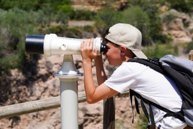 Bir çocuk teleskopla bir dağa bakıyor. Çocuk beyaz bir gömlek ve şapka giyiyor.